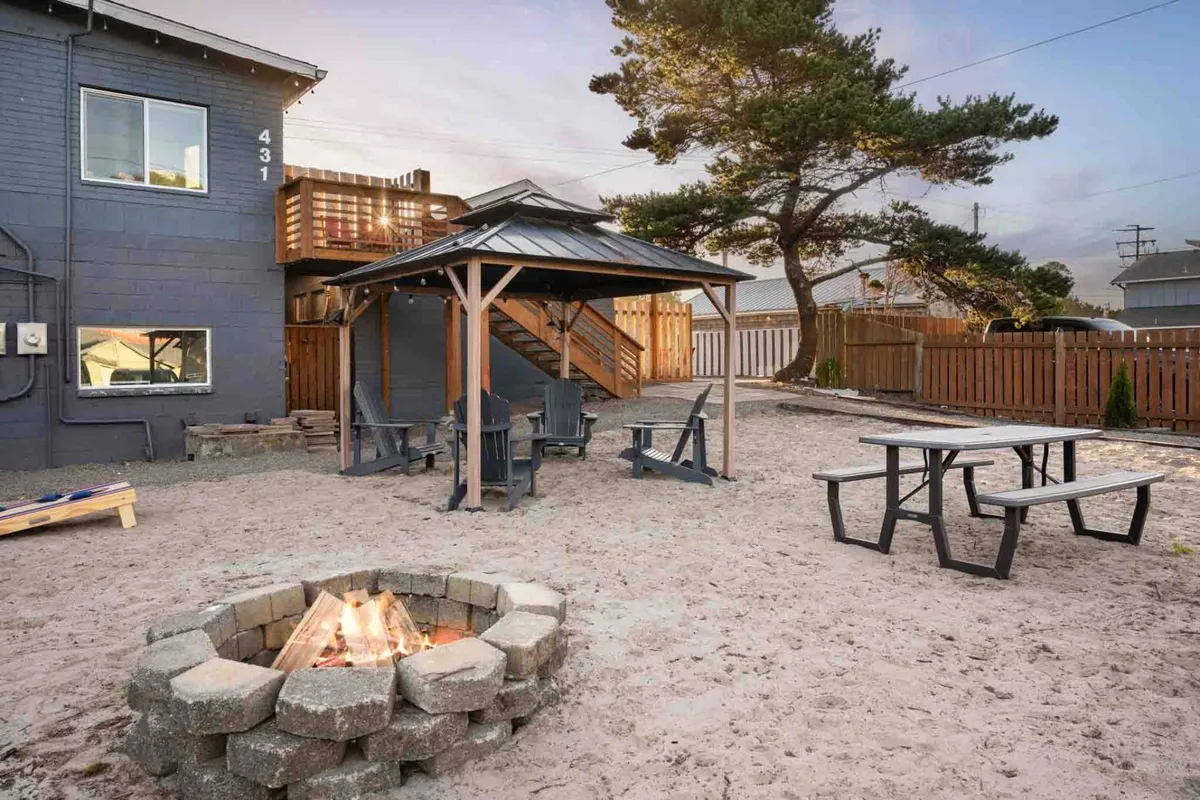 A spacious gazebo area with a fire pit, Adirondack chairs, and outdoor seating arranged on a wooden deck. The gazebo is set for socializing, with the warm glow of the fire inviting guests to gather and enjoy the sunset or evening sky.