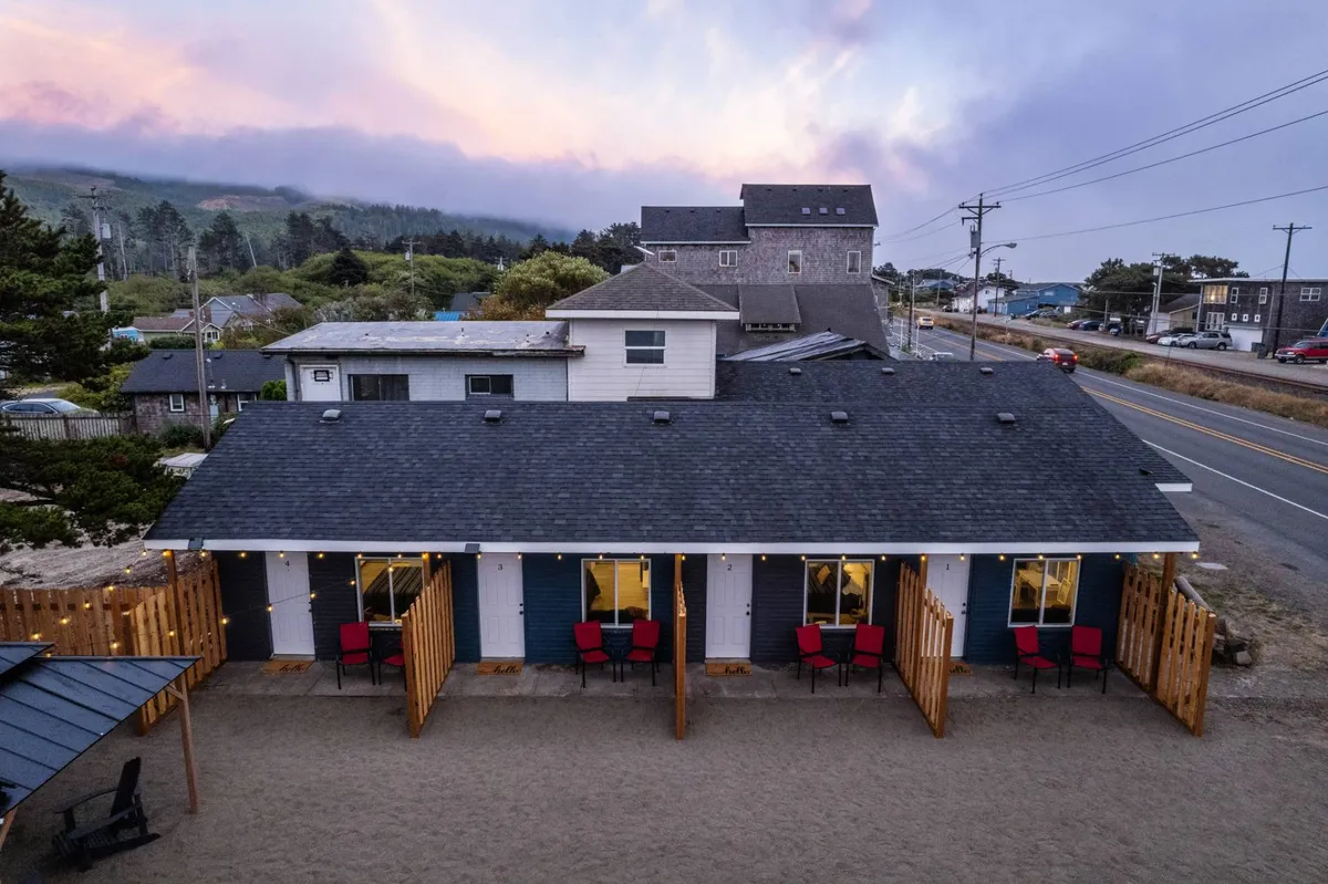This image showcases a picturesque row of cottage-style accommodations, each with inviting red chairs on the porch. The sandy area in front of the cottages and the scenic hills and clouds in the distance evoke a tranquil, welcoming atmosphere perfect for relaxing and socializing outdoors.