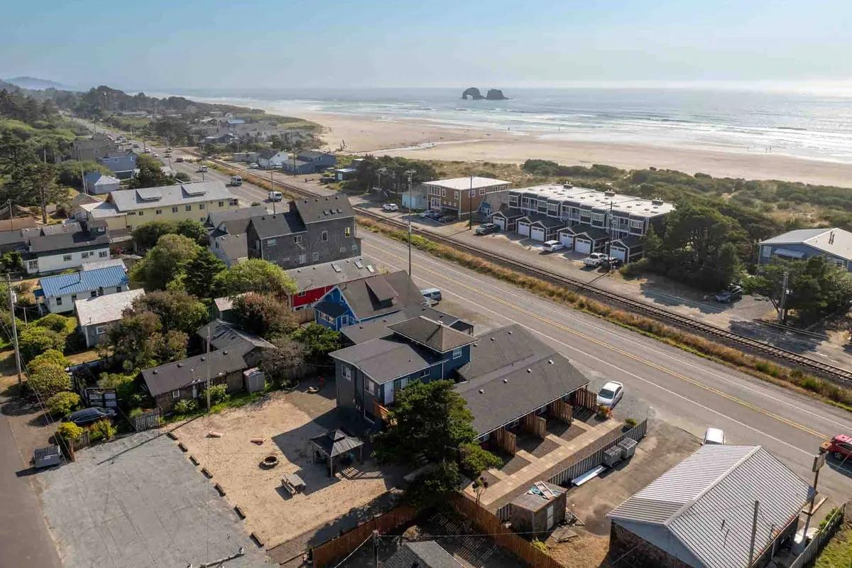 A relaxed vista showcasing the sandy beach, calm ocean, a road, and nearby houses. The scene feels welcoming and lively, hinting at easy access to the water, as well as the community vibe of the neighborhood. People can be imagined strolling to the surf or taking part in local gatherings.