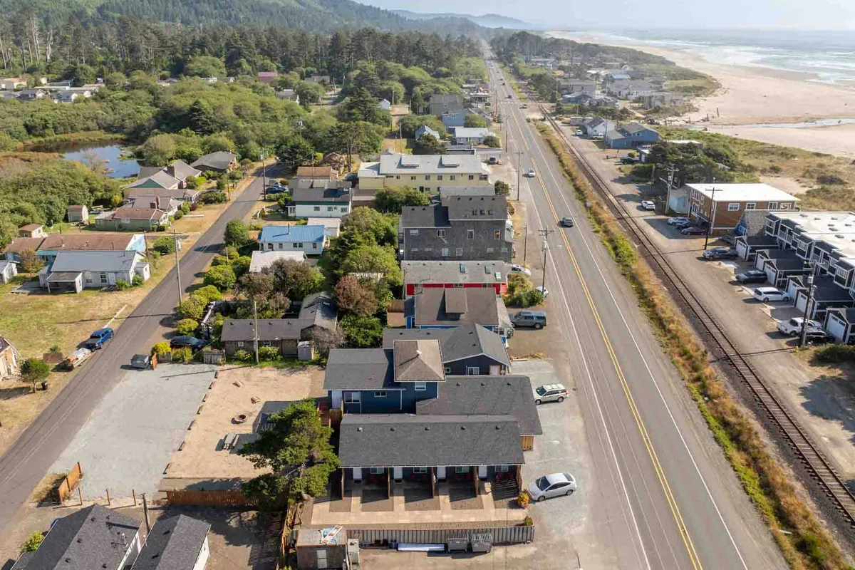 A wide exterior shot featuring rows of charming cottages, green spaces, a main road, and direct access points to the beach. The image conveys a sense of openness, community, and immediate entry to coastal fun.