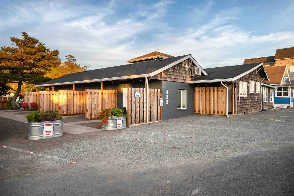 A welcoming exterior view of one of the cottages, bordered by wooden fences and vibrant flowers, with the shingle exterior and house number adding charm. The porch offers a cozy spot to relax, and the sunset lighting enhances the romantic appeal.