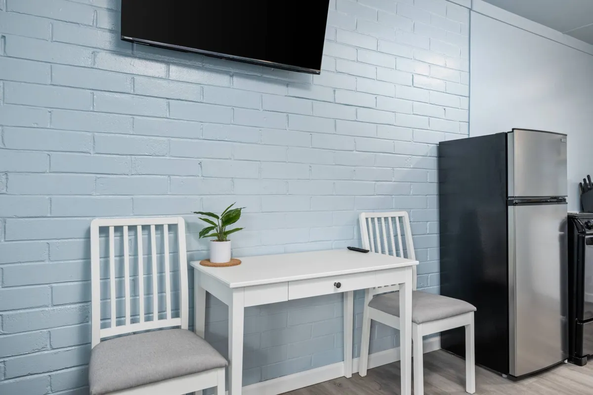 A cozy dining area with a white table, two chairs, a green plant centerpiece, a TV in the background, and a black refrigerator nearby. The setting is inviting and functional, perfect for casual meals or relaxed mornings.