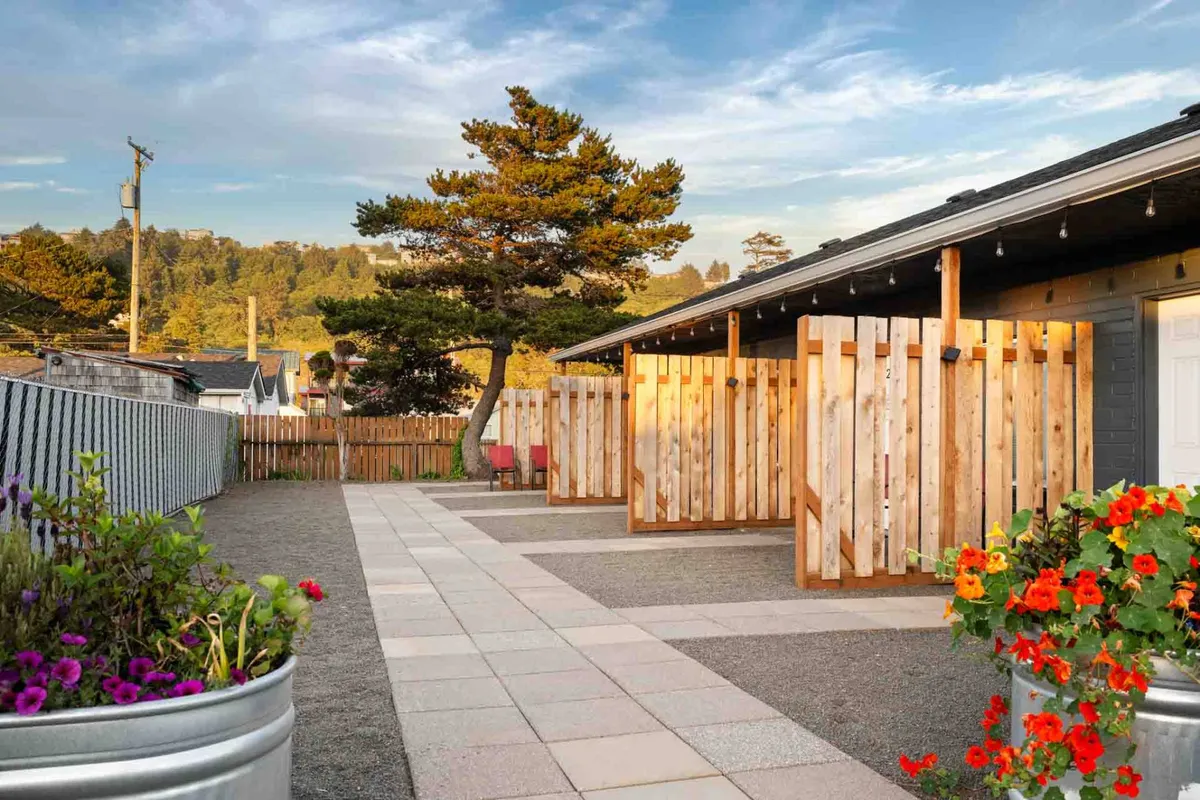 A cheerful exterior scene with wooden fencing, vibrant flower pots, and a tidy pathway leading up to private entrances. The small patios provide a private relaxation area perfect for both guests and their pets.