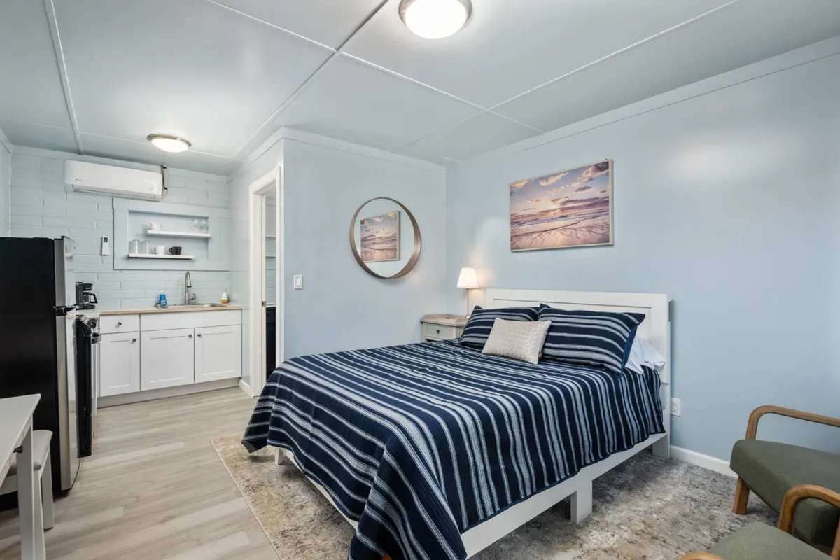 A warmly lit cottage bedroom featuring a striped bedspread, a round mirror above the bed, framed beach-themed artwork, and a compact kitchenette. Sunlight streams through the window, casting a gentle glow on the inviting furnishings and light-toned decor.