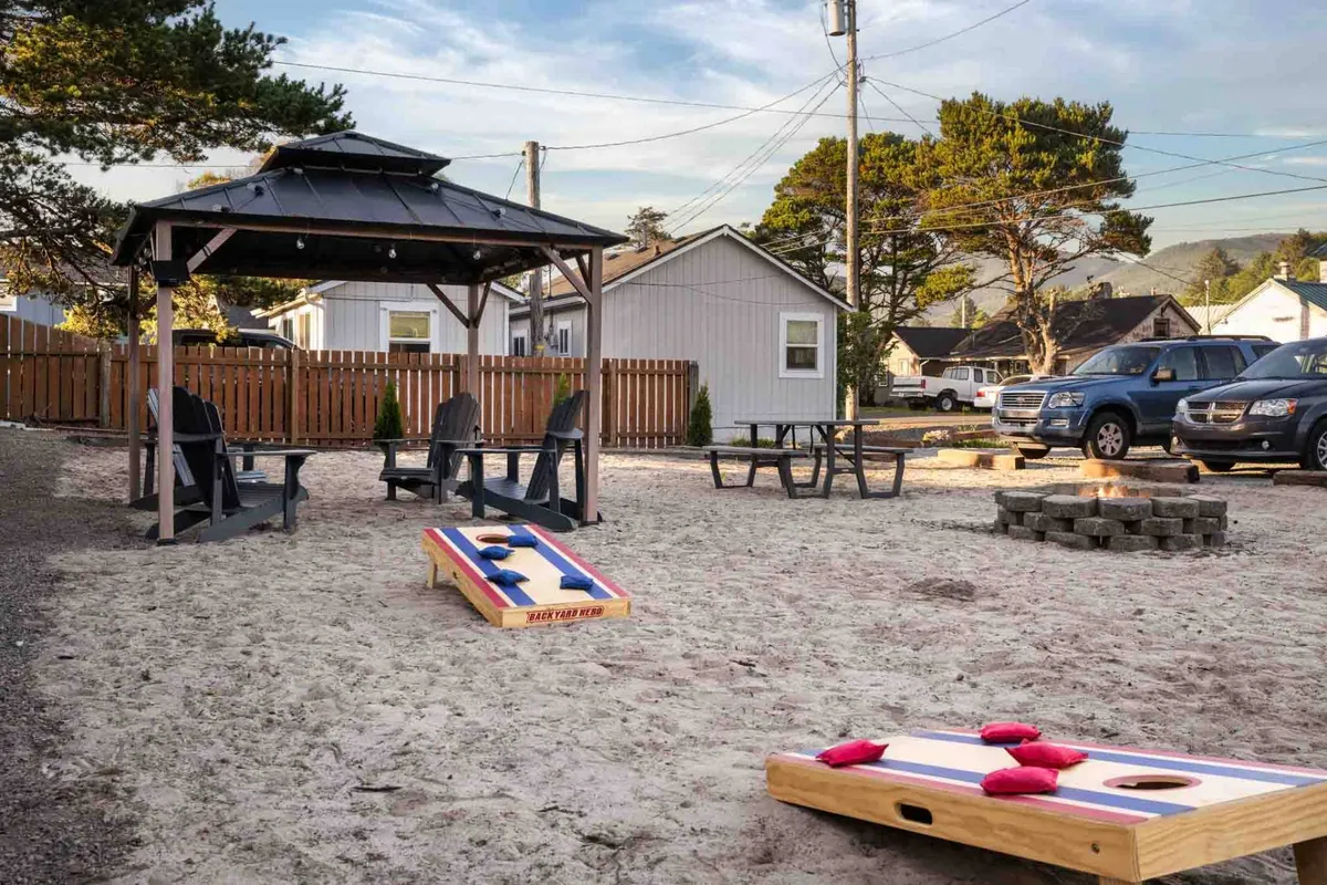 A welcoming outdoor communal area featuring a gazebo, several outdoor seating options, a cornhole game, and a fire pit. The space is set against a backdrop of lush greenery, offering a pet-friendly place for relaxation and socializing.