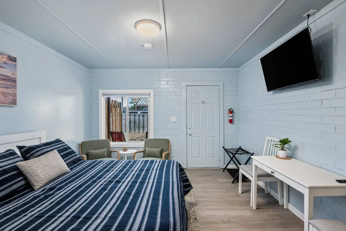The image showcases a bright, freshly renovated condo interior with a stylish striped-bedding bed, a cozy seating area perfect for morning coffee, a large wall-mounted TV for relaxing evenings, and a mirror that fills the space with natural light, all coming together to create a comfortable and inviting atmosphere.