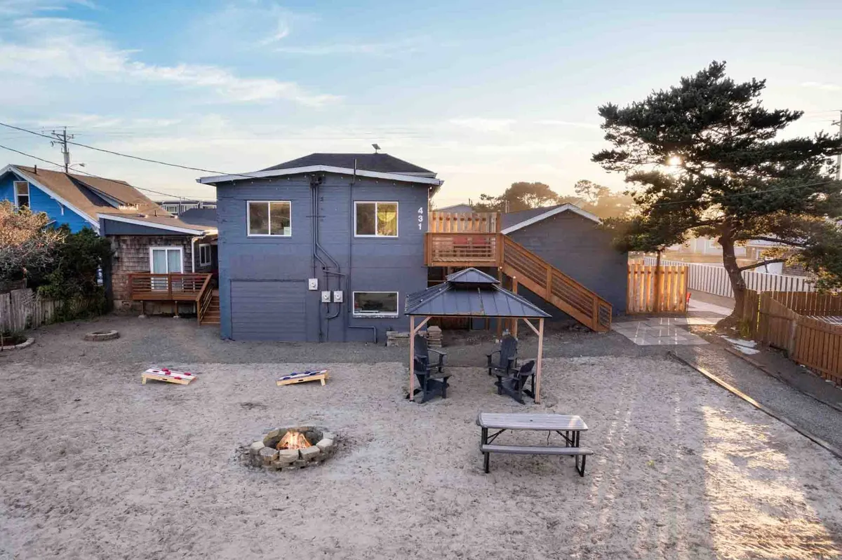 This image captures a lively outdoor communal area featuring a fire pit, gazebo, picnic table, upper deck, and a sand play area. The space is inviting, with multiple cozy spots for relaxing, sharing meals, or playing games, all set in a cottage-style landscape.