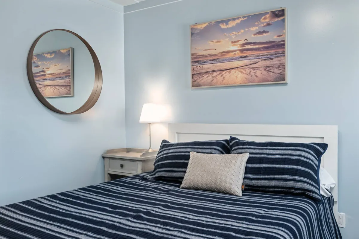 A tranquil bedroom with a comfortable bed adorned in striped bedding, a bedside lamp casting soft light, and a framed photo of a beach sunset on the wall. The room feels serene, inviting you to unwind and relax.
