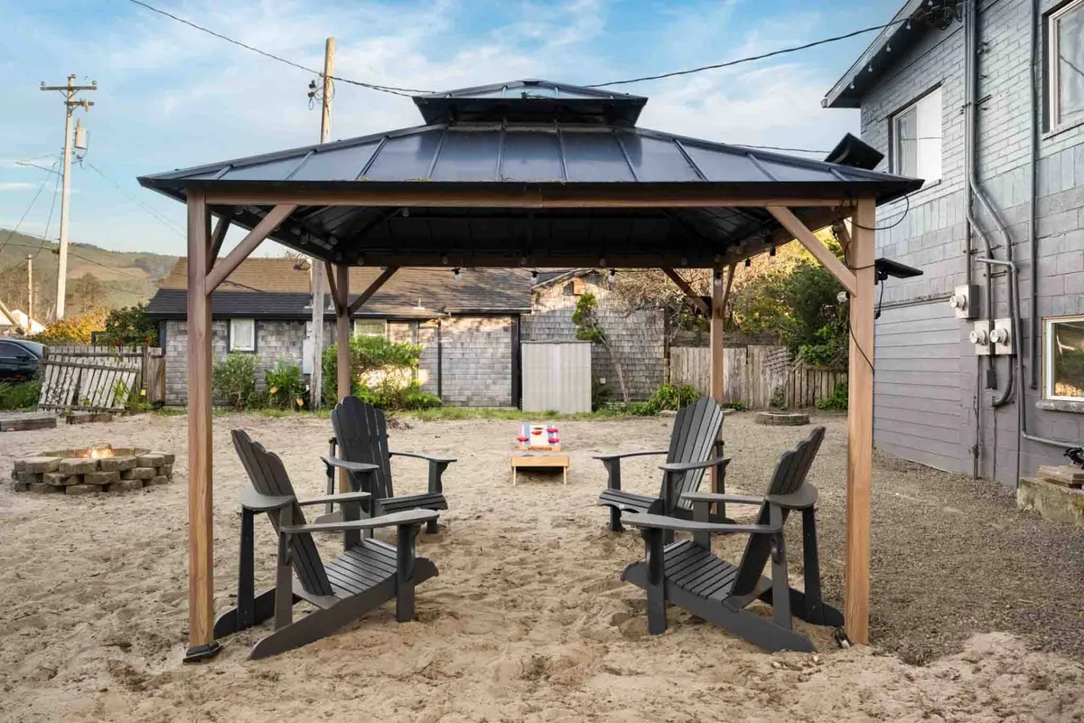 A communal outdoor space featuring a gazebo, Adirondack chairs circling a fire pit, and wooden decking. The scene is casual and inviting, hinting at friendly gatherings, stargazing, and relaxation under the open sky.